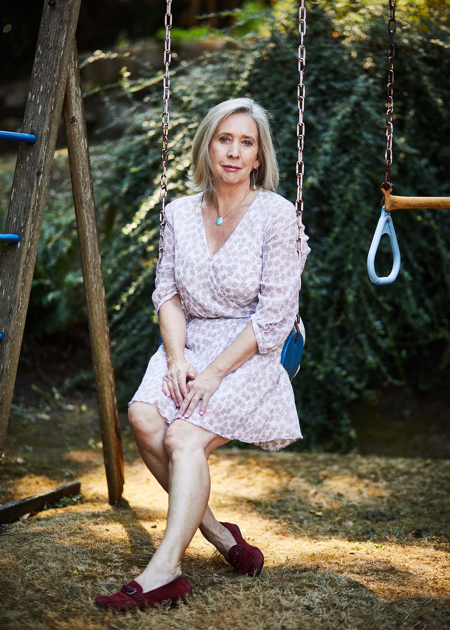 Photo of Warren Binford, wearing a white and pink dress with a spotted pattern, sitting on a kid's swing set outside in someone's back yard. She smiles slightly, a bit somber, and has blonde shoulder-length hair.