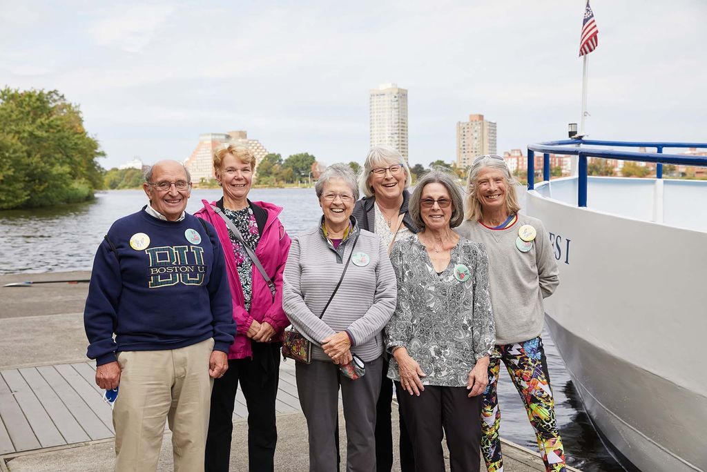 Photo taken during the Charles River for Golden Terriers during Alumni weekend. A group of six gray-haired alumn smile as they stand on a dock along the Charles.