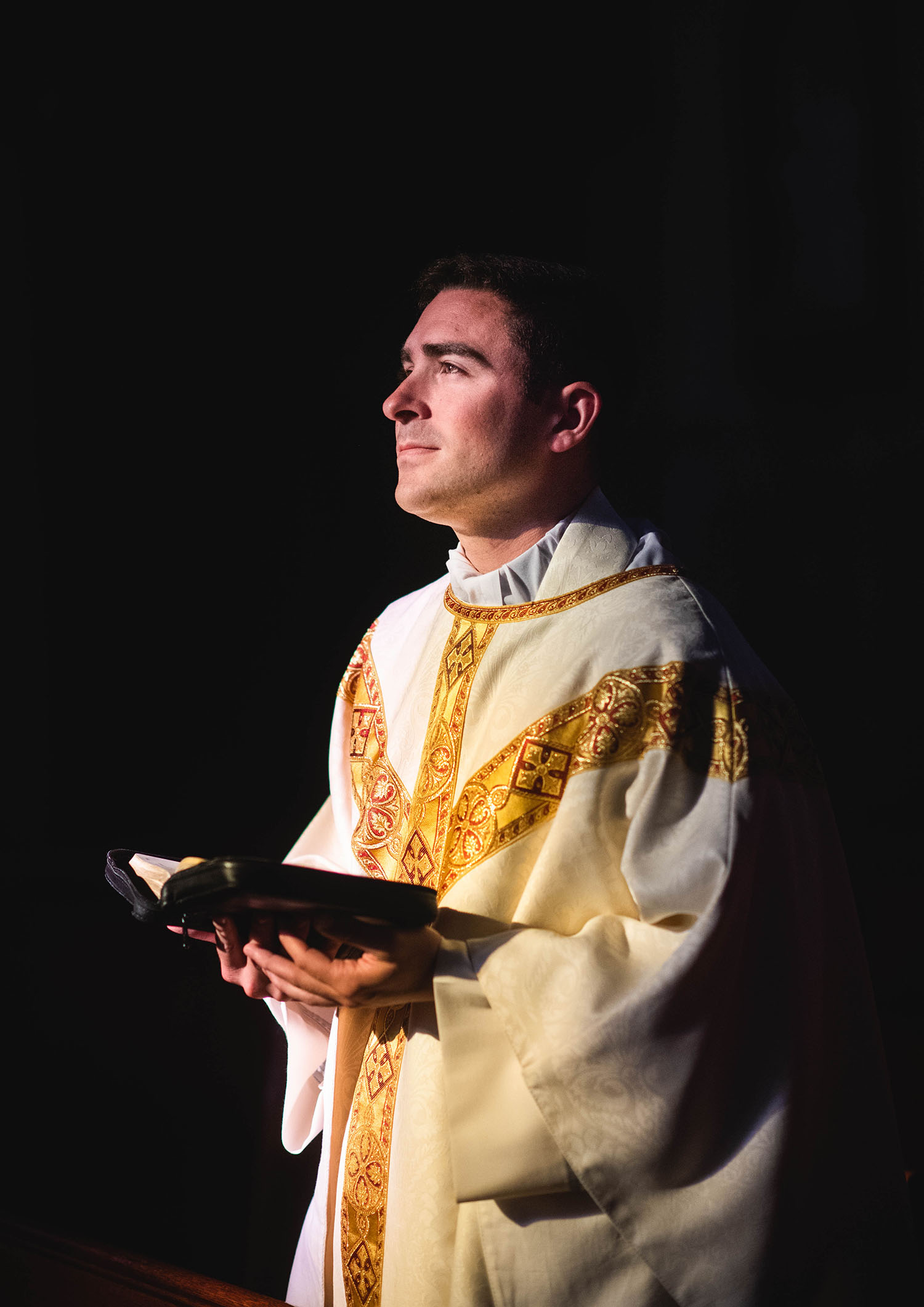 Photo of The Rev. Robert LeBlanc (CGS’11, COM’13) celebrates mass at St. Mary of the Assumption in Brookline, Mass.. He hold a bible in his hands and looks of towards his right into the middle distance. He wears a white robe with gold trim and has short brown hair. The background is black.