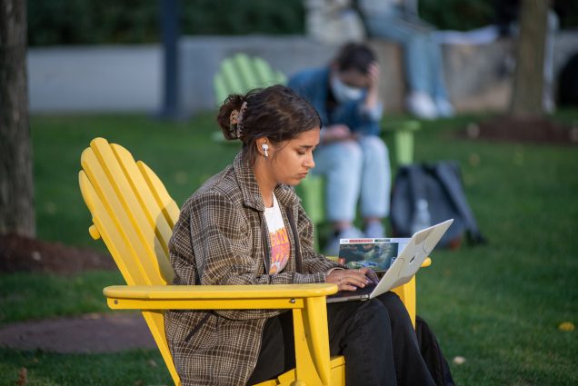 Photo: Amanda Cintron (COM’32) working on mythology essay at COM Lawn Oct 12. A student swearing a white tee and oversized plaid collared shirt sits in a large yellow chair on a green lawn working on their laptop. Other students sitting in similar large chairs are seen behind them in the blurry background.