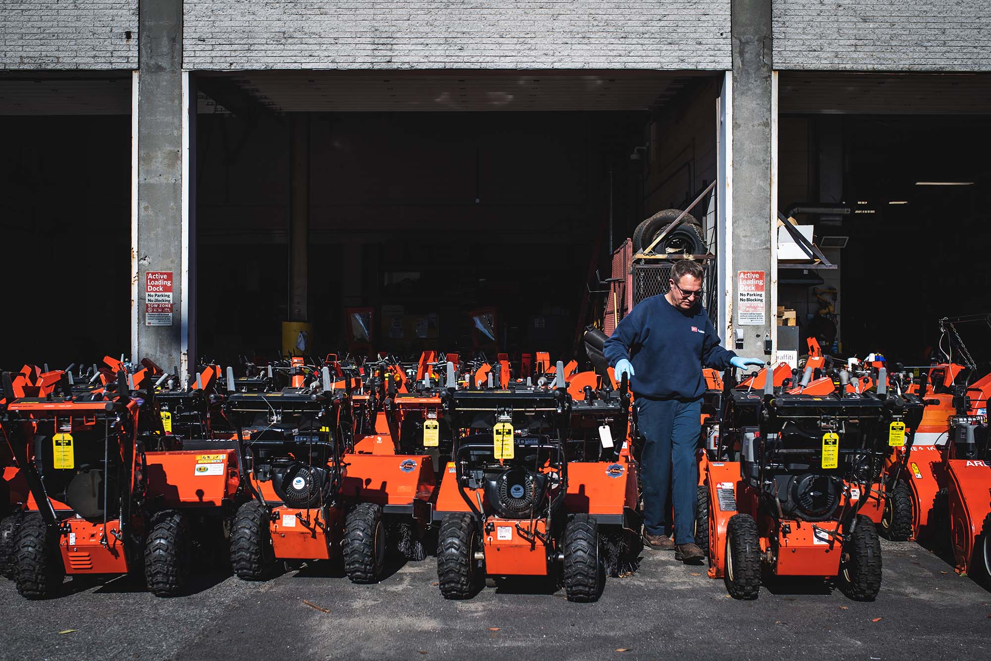 Photo: Craig Espenhain of BU Campus Planning and Operations walks through rows of snowblowers on October 19, 2022. A man in a navy blue sweat suit walks amongst rows of bright orange snowblowers.