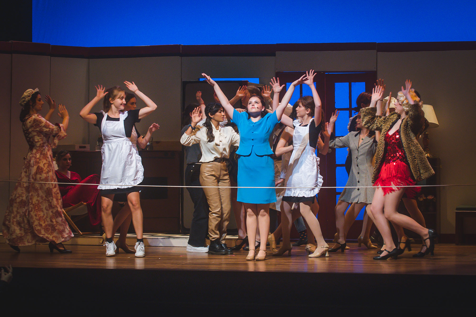 Photo: Students raise their hands on stage as they perform a musical number. The lead actor wears a blue blazer and skirt combo, as people wearing various outfits ranging from a maid's uniform to a sequin red dress raise their hands around her on stage.