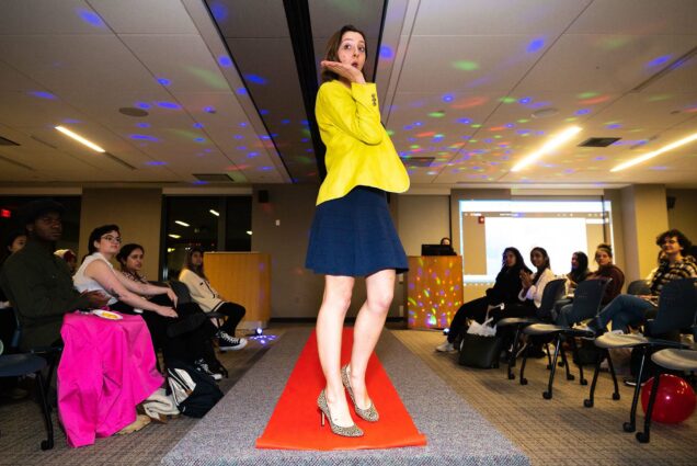 Photo: Fiona Schicho (School of Social Work ’25) blows a kiss after walking down the runway during the fashion show at the Professional Clothing Closet’s Nov. 16 event. A young white woman wearing a bright yellow blazer and navy, knee length skirt, poses and blows a kiss in the middle of a fashion runway lined with red carpet. The audience sits on either side of said runway.