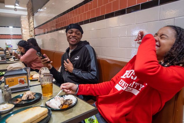 Photo: At West Dining Hall, Toby Makoyawo (CAS’25), left, enjoys a turkey dinner and good company with friends Jada Smith (CAS’26), at left, and Matthew Spicer (CAS’25), not pictured, and Ijeoma Ezechukwa (CAS’26). On various plates around the table are the traditional Thanksgiving meal complete with turkey, local mashed potatoes, stuffing, sweet potatoes, cranberry sauce, and pumpkin pie. A young black man wearing a black durag and black and grey hoodie laughs with cellphone in hand as a young Black woman to his left, wearing a red BU track and field hoodie, leans back with laughter.