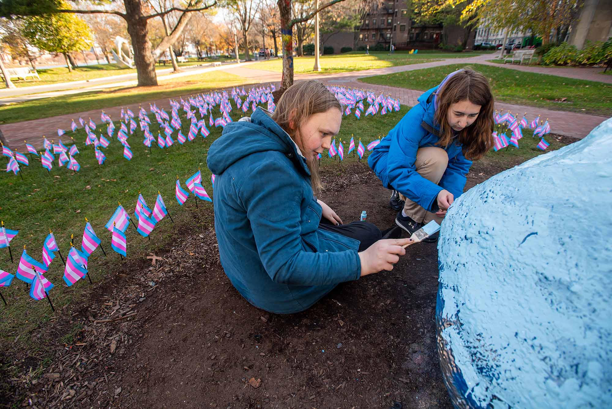 Photo: VP of Trans Listening Circle Lena Broach (CFA’25), left, and President of Trans Listening Circle Felix Wheeler (COM’23) paint the rock in the colors of the transgender flag after placing flags on Alpert Mall Nov 18 to honor the lives of trans and gender-diverse people reported murdered worldwide in the past twelve months. Two young women wearing blue, puffy coats paint a rock. Behind them, small, transgender flags are lined and placed into the lawn.