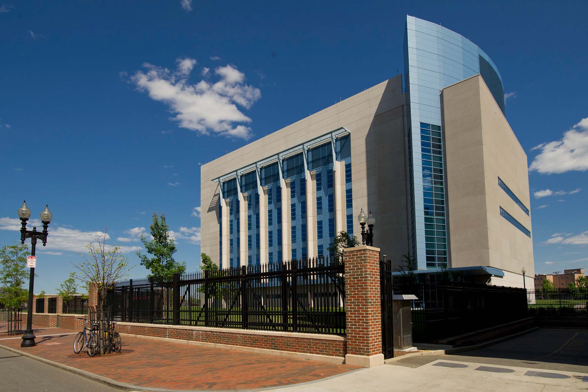 Photo: Exterior of the NEIDL building. A large, beige building with blue windows is shown behind an iron fence on a sunny day.