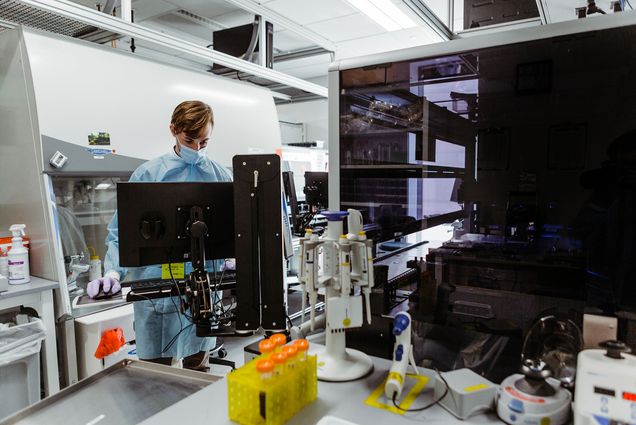 Photo: The oldCovid-19 testing lab at Kilachand Center. A researcher wearing a light blue mask and gown works behind a large computer as machines and vials are shown on the counters in front of them.