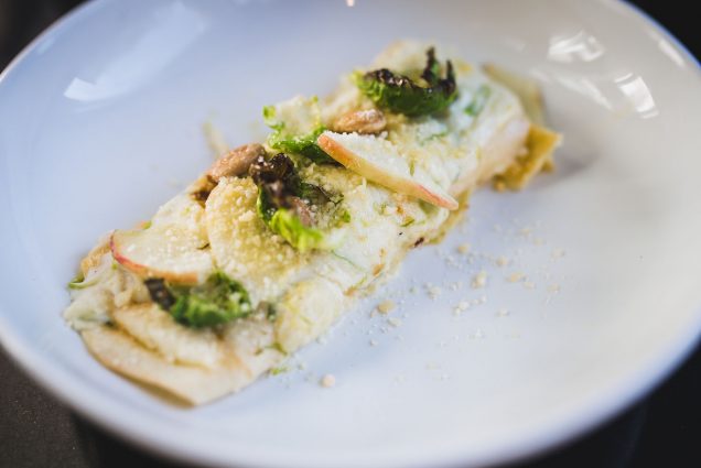 Photo: A large piece of butternut squash manicotti topped with Brussels sprouts, pickled apple, and almonds is shown on a white plate. A slight sprinkling of Parmesan cheese tops the dish.