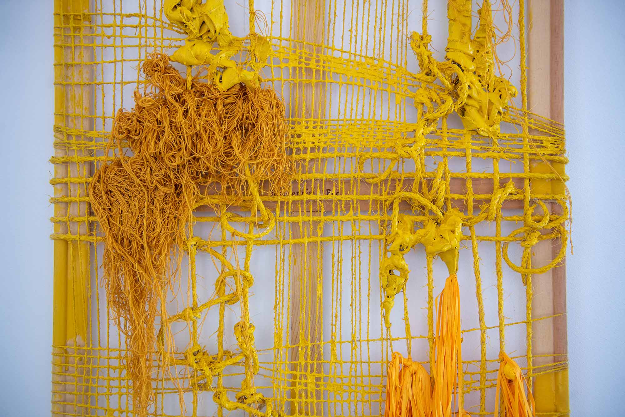 Photo: A large tapestry hangs on a white gallery wall. Tapestry is made up of various shades of yellow threads and materials in various criss-cross patterns.