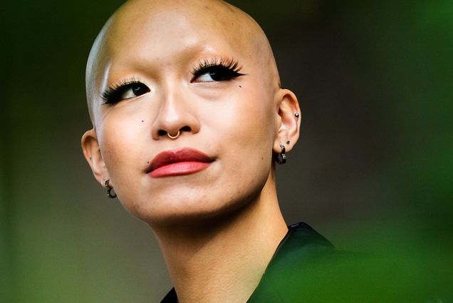 Photo: Katarina Quach poses for a photo on November 15, 2022. A young, bald, tan woman with long lashes and wearing red lipstick and a large black, oversized blazer, poses for a photo in front of a dark backdrop. Blurred, green leaves in the foreground at dimension to the photo.