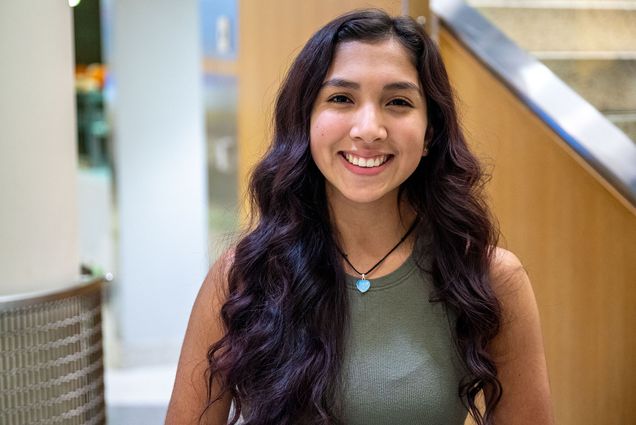 Photo: Madison Miranda. a young Puerto Rican woman wearing a cropped olive green tank and jeans, smiles and poses next to a wooden staircase.