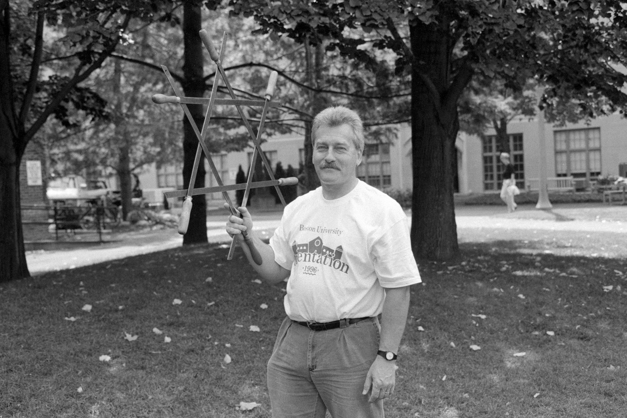 Black and white photo of Anthony Barrand. A white man wearing a white tee shirt that reads "Boston University Orientation 1996" tucked into slacks holds up in one hand a large, wooden jewish star. He stands on a BU lawn on campus.