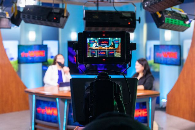 A photo of a TV camera and camera screen in focus are in the center of the image, showing two student news anchors behind a news desk with The Wire graphics on a screen behind them. The two students are out of focus on either side of the camera, showing the rest of the TV studio, with big lights suspended from the ceiling above.