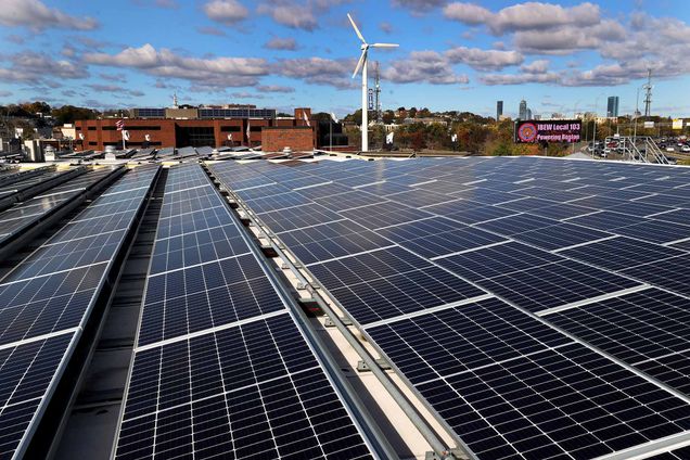 Photo: Newly installed Nexamp solar panels at the Local 103 headquarters in Dorchester. Large solar panels cover a rooftop on a Boston building. A city skyline and distant white wind turbine are shown in the distance.