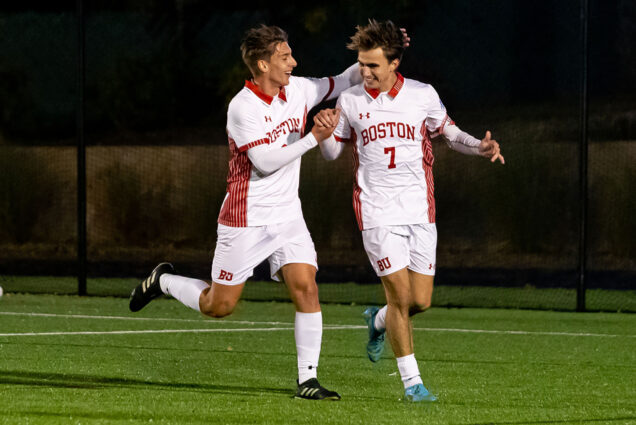 Photo: BU men’s soccer players Gianluca Arlotti (CAS’23) (left) and Quinn Matilus (CAS’23) celebrating a goal against Lehigh October 29. Two young men wearing white and red soccer uniforms grip hands and raise their other arms in cheer as they smile and run down the field.