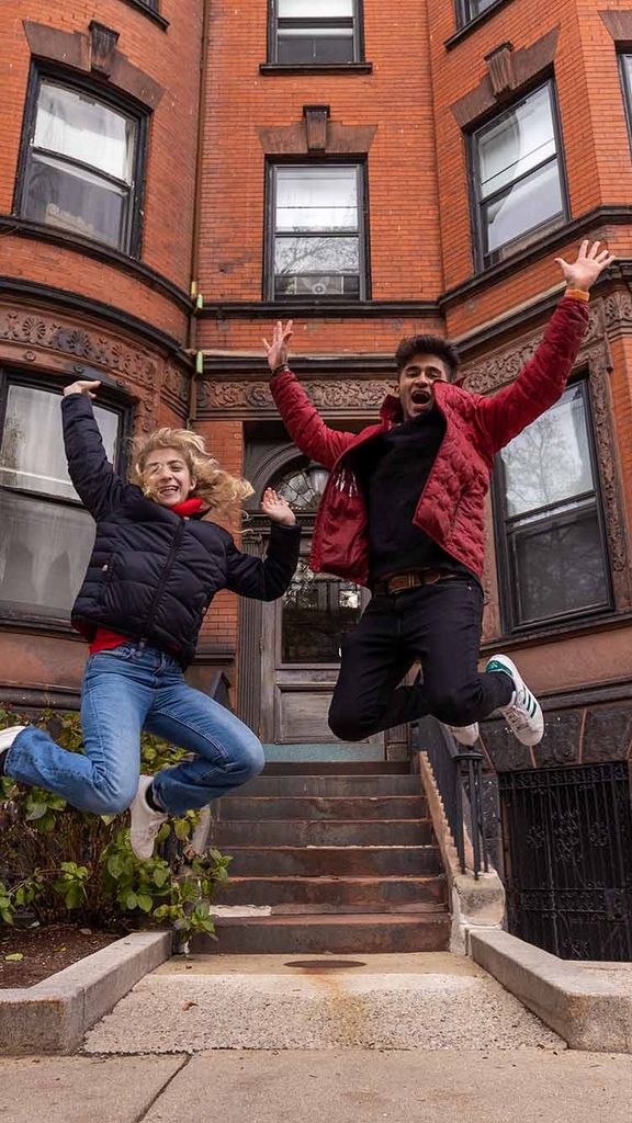 Photo: Sophia Delia (ENG’23) (left) and Rohin Bajaj (ENG’25) are caught mid-jump in front of a red, brick brownstone building. A young white woman and a tan young man both jump and laugh on the sidewalk in front of a brownstone. Both wear puffy winter jackets.