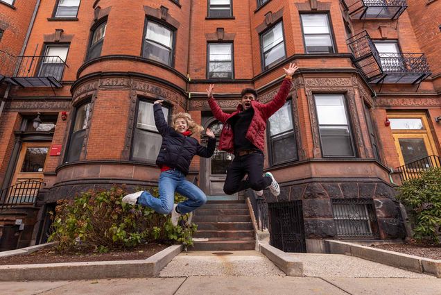 Photo: Sophia Delia (ENG’23) (left) and Rohin Bajaj (ENG’25) are caught mid-jump in front of a red, brick brownstone building. A young white woman and a tan young man both jump and laugh on the sidewalk in front of a brownstone. Both wear puffy winter jackets.