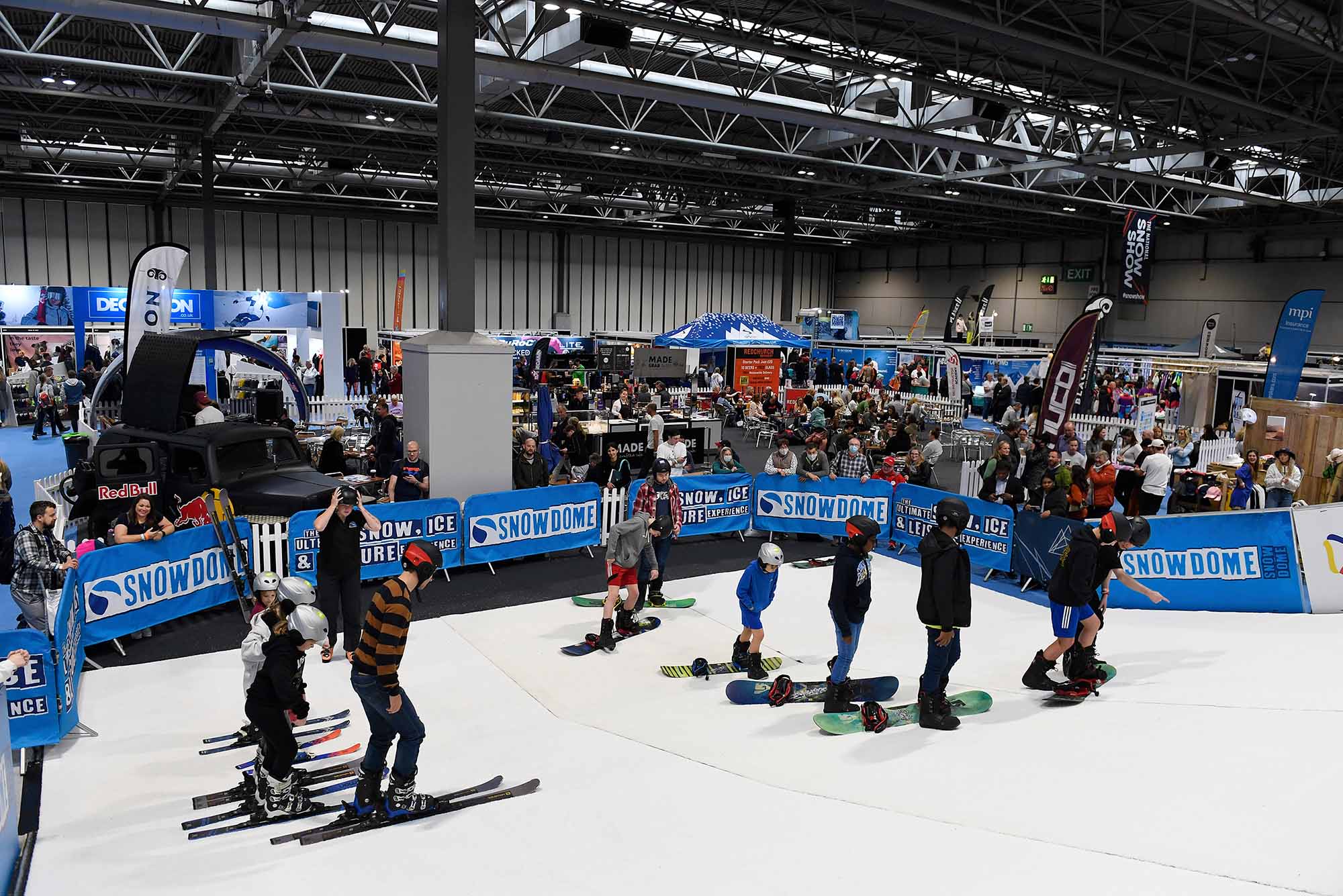 Photo: A group of people use snowboards on a small patch of snow in a convention center. Convention goers and booths are on the outskirts of the snow center.