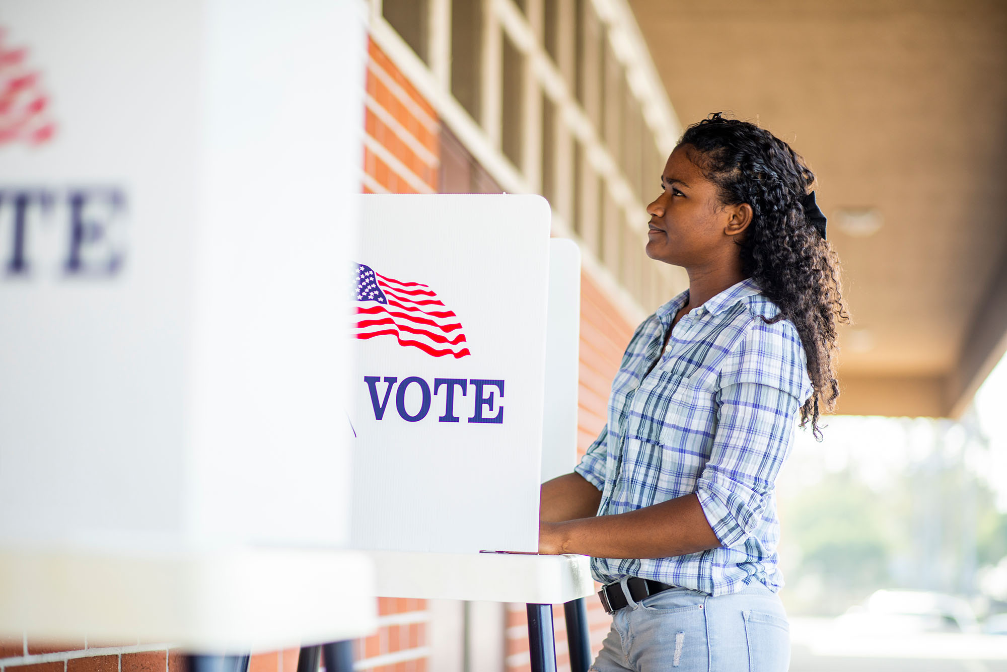 Photo of a young black girl voting on election day. A young black girl with curly hair votes in a voting booth set up outside a brick building.