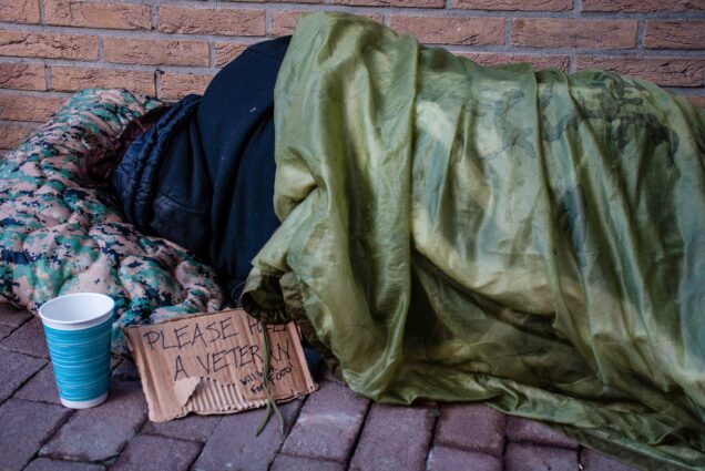 Photo: A homeless veteran sleeps on the ground with their back facing the camera outside of a brick building with a sign begging for change.