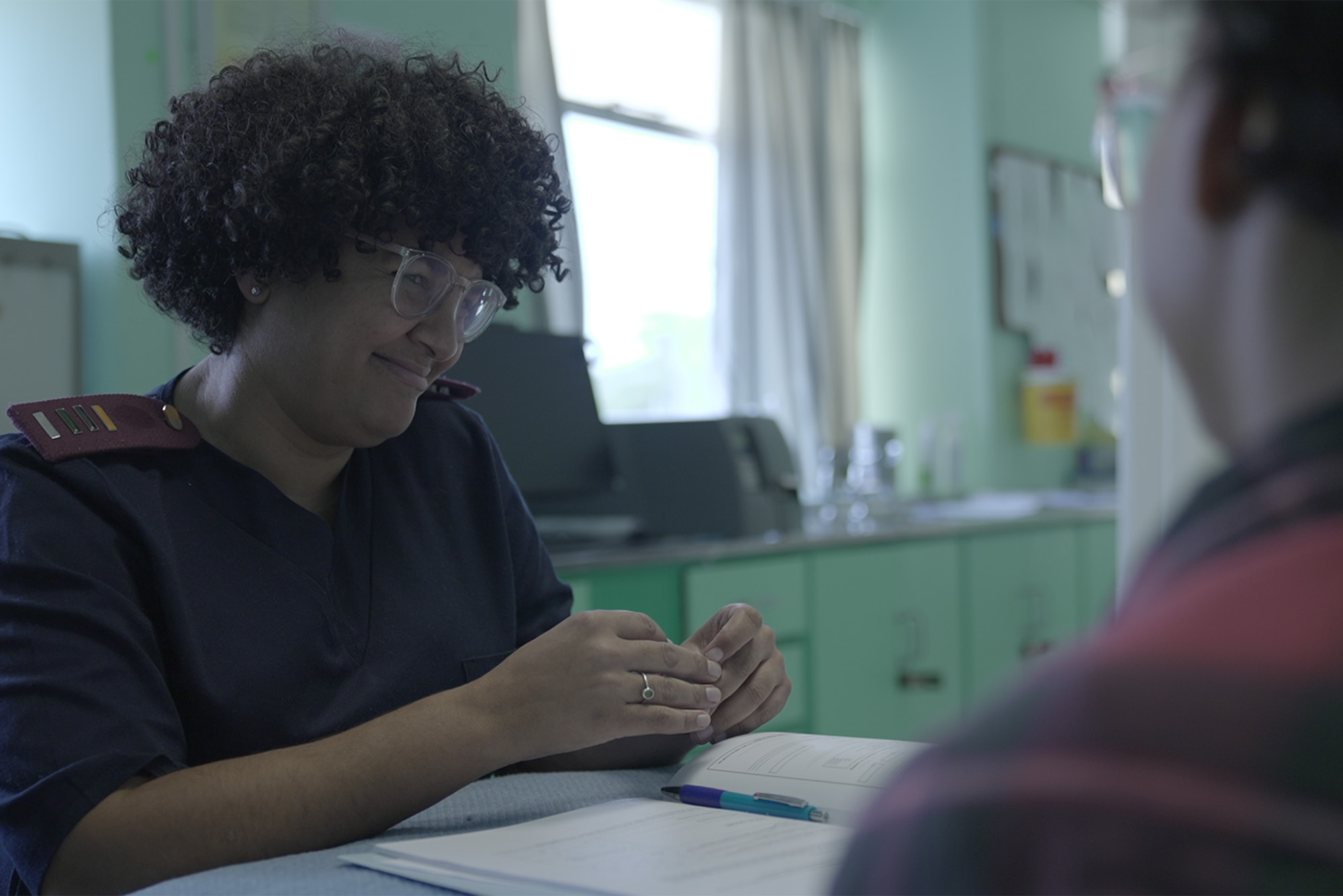 Movie screenshot: A black woman with short, curly hair and wearing navy scrubs sits and smiles at a patient. In the blurry foreground the back of the patient can be seen sitting across from the nurse.