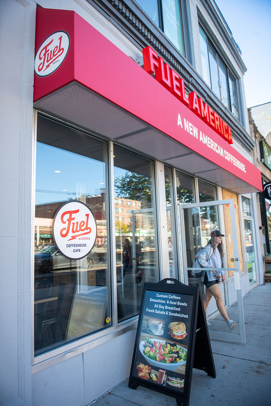 Photo: Exterior of Fuel America. Large red letters read "Fuel America" as the red canopy below reads "A new American Coffeehouse" both over the entrance for the shop. A person is seen exiting the shop and a small billboard in front advertises some treats.