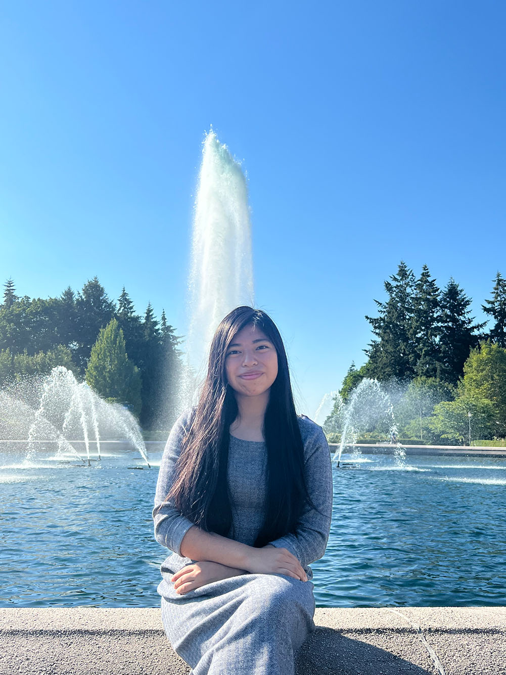 Photo: Teresa Miguel sits and poses for the camera in front of a large water fountain. A Hispanic young woman wearing a long grey maxi dress smiles for the camera.