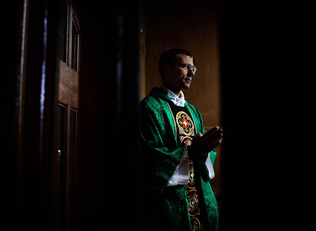 Photo of Rev. Michael Zimmerman with his hands clasped in prayer, leaning against a wooden wall. He is lit, but the background is almost black.