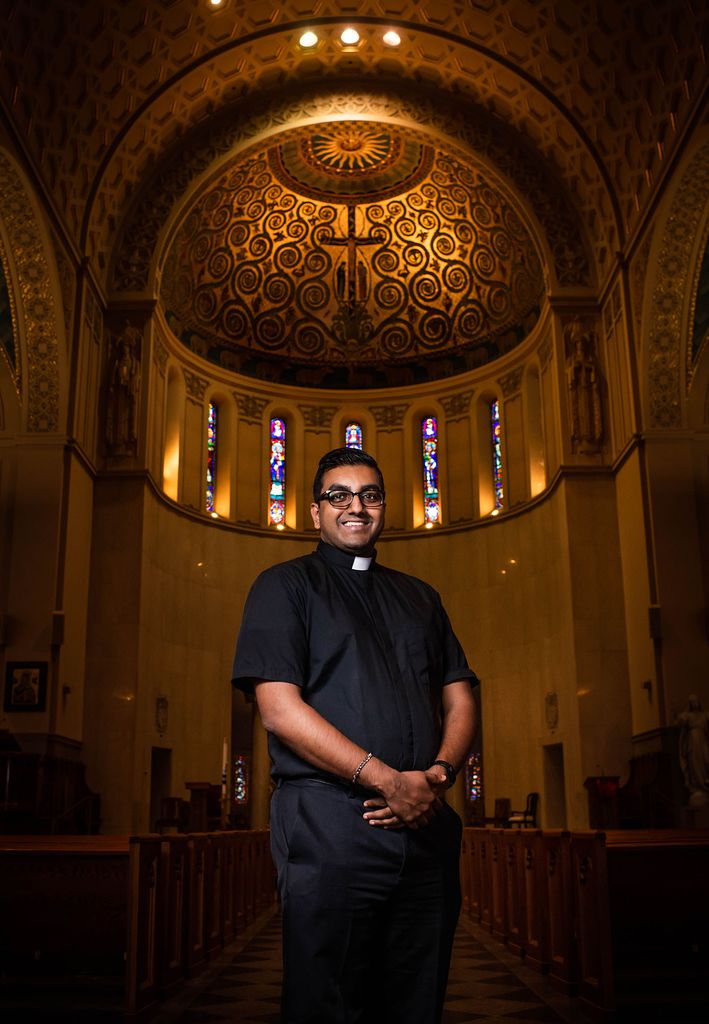 Photo of Fev. Denis Nakkeeran, standing in front of dome with his hands clasped. He smiles, has black glasses, and olive skin, and wears a black priest's outfit. The dome behind him is lit and looks golden.