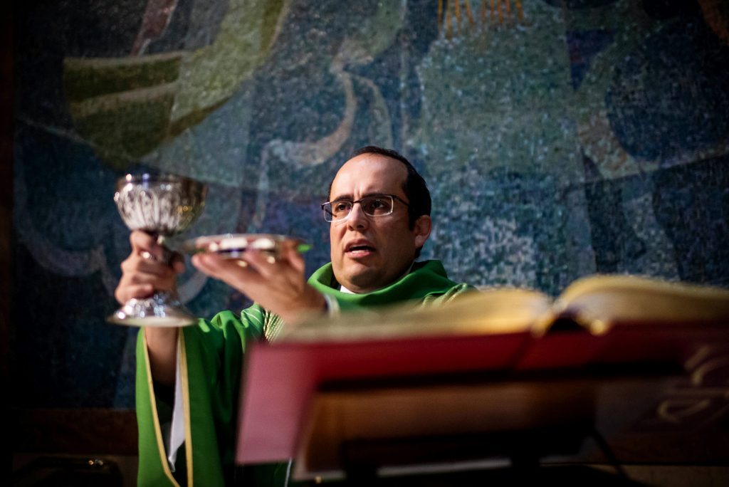 Photo of The Rev. Carlos Suarez (CGS’01, CAS’03) (below), pastor of St. James & Immaculate Conception in Stoughton, Mass, holding a silver chalice and small plate. He stands behind the altar, and an open religious text is seen in the foreground. Behind him is an intricate mosaic.