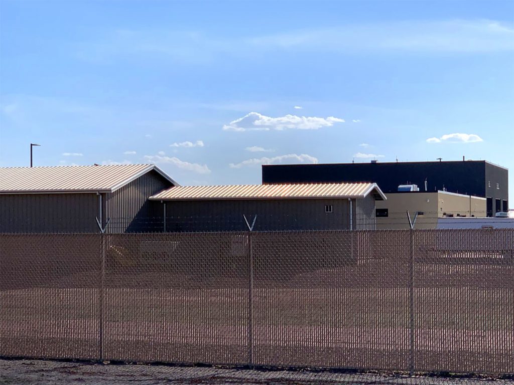 Photo of the Clint Border Patrol facility taken in 2019. The facility is hidden behind a metal fence. Blue sky is seen behind it. 