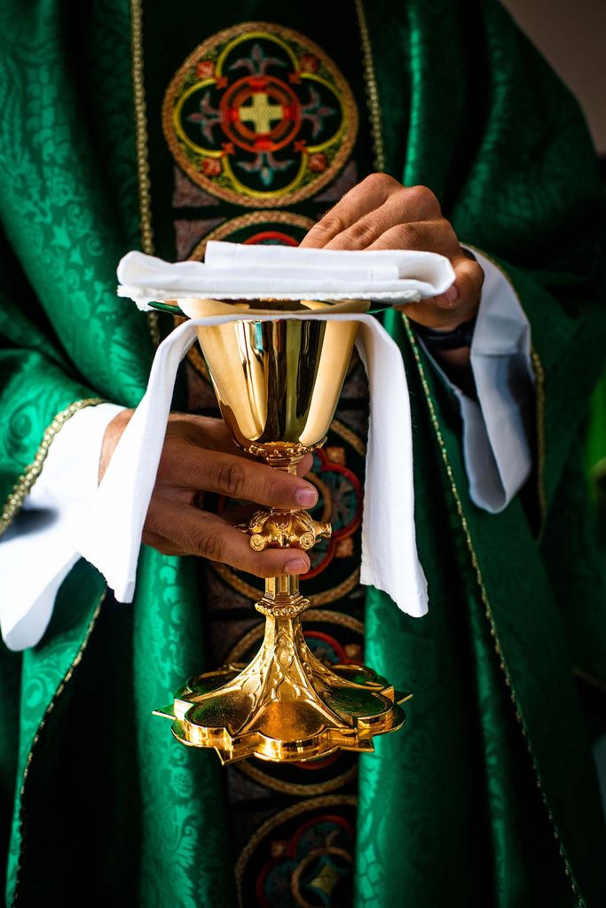 Photo of a priests in a green robe holding a gold chalice with a white cloth folded over it.