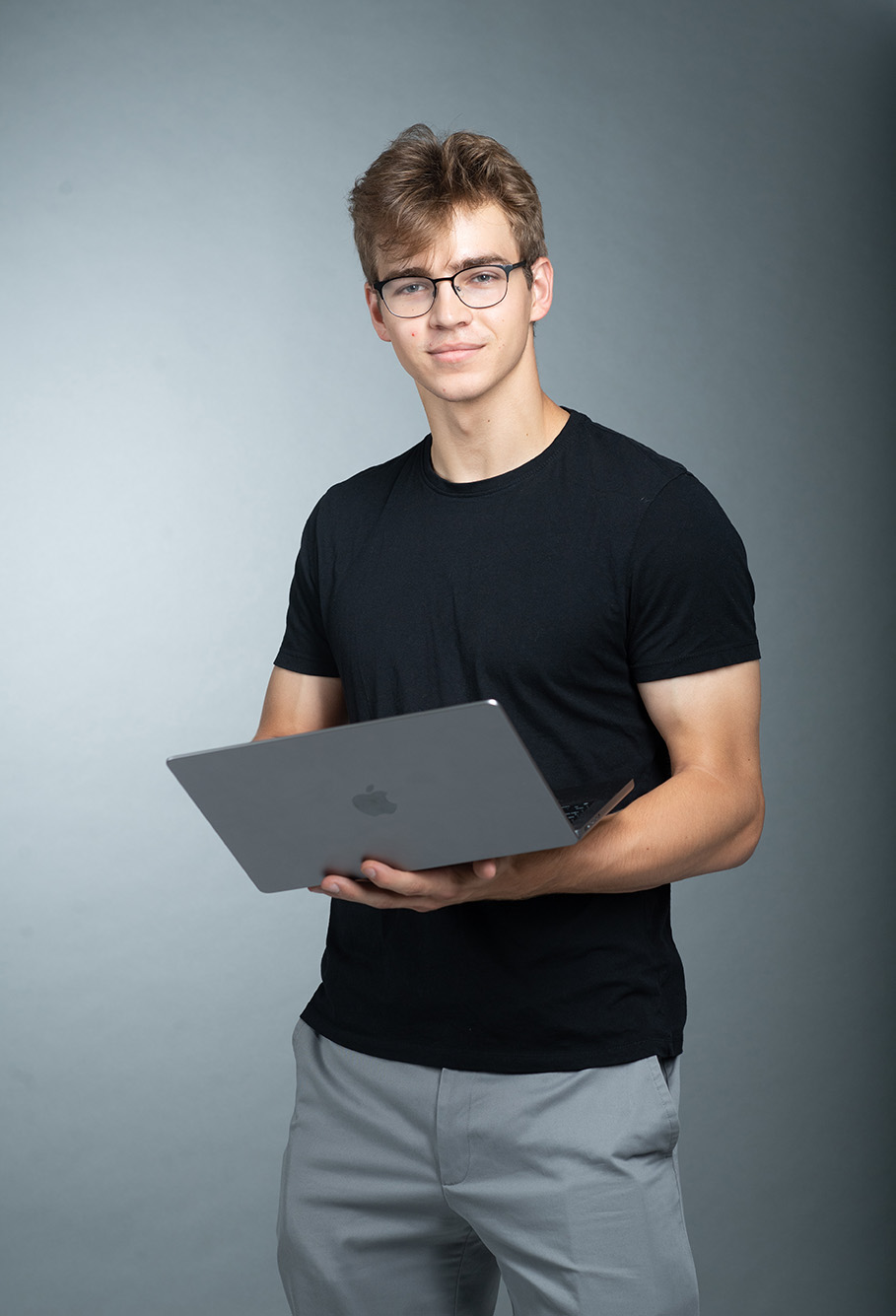 Photo: Ben Gardiner poses in front of dark grey backdrop with Apple laptop in hands. A young white man with wearing glasses, a black short-sleeved shirt and greypants, stands and poses with his laptop in held in front of him.