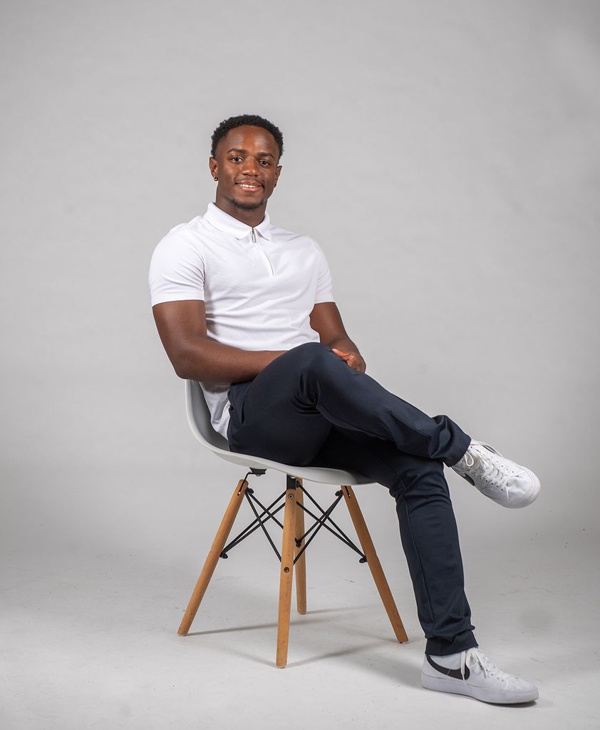 Photo: Louie Belile, a young Black mean wearing a white polo shirt and jeans, sits and poses with legs crossed in front of a grey backdrop.