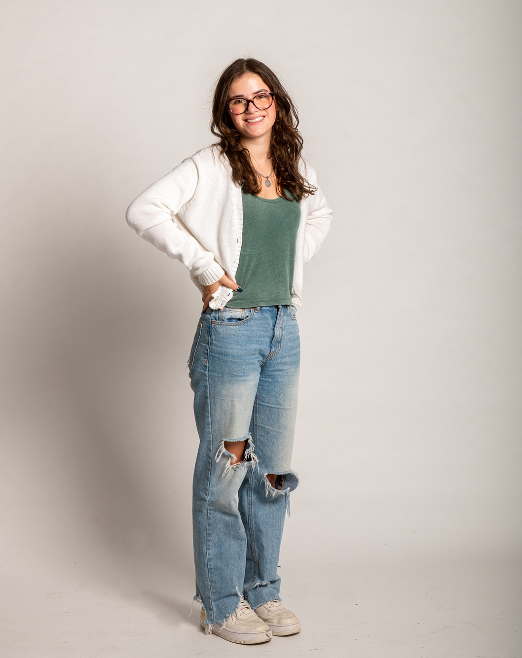 Photo: Elizabeth Rosen, a young white woman wearing glasses, a green blouse, white cardigan, and ripped jeans, stands with hands on hips and poses in front of a beige backdrop.