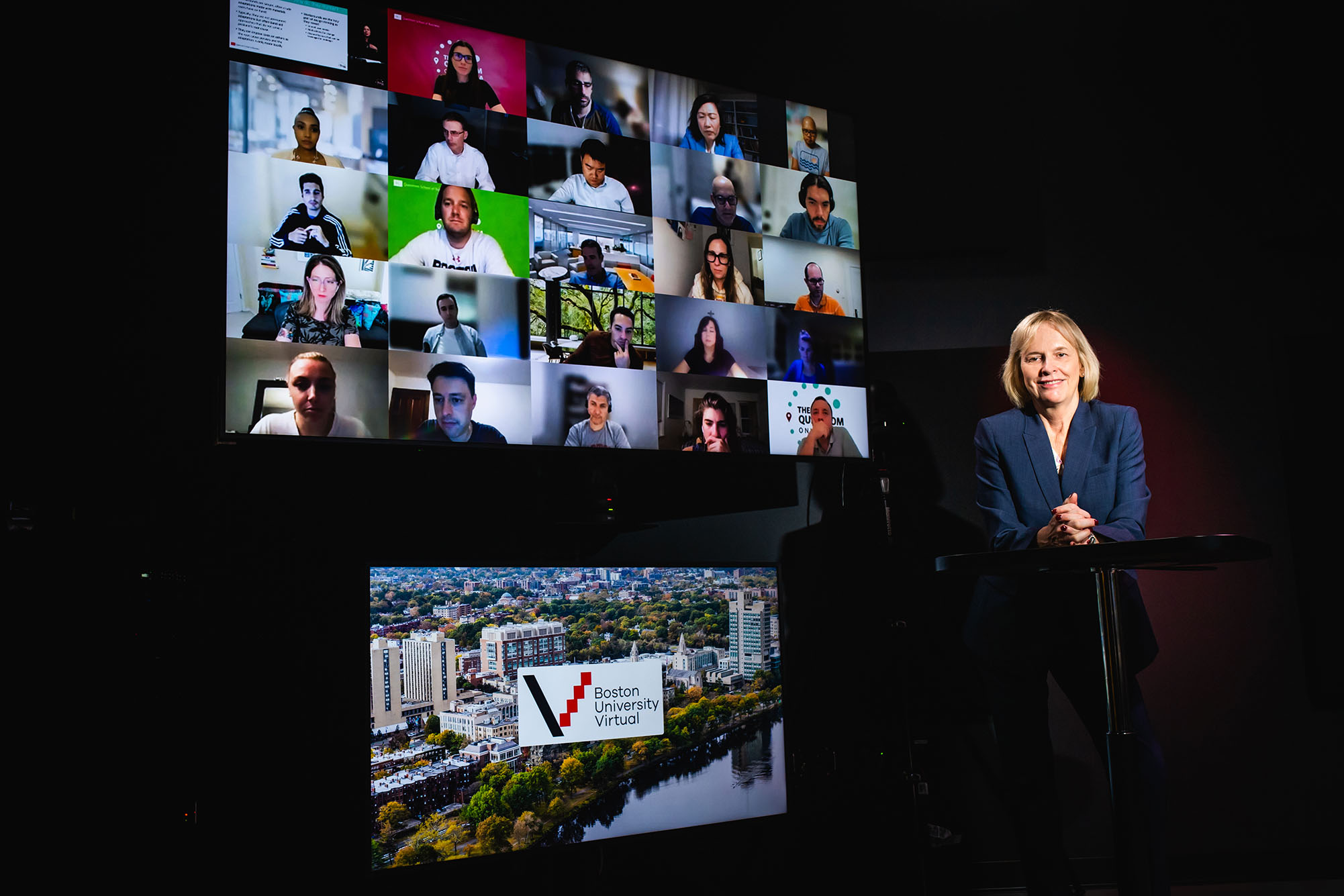 Photo: Wendy Colby, BU's first vice president and associate provost for BU Virtual, poses for a photo in a Questrom online studio. A white woman with shoulder length blonde hair and wearing a navy suit ensemble sits in a chair with hands folded over crossed legs. She sits in a dark room lit with purple backlights as two large monitors are shown to her left. The top displays a grid of students Zooming in viewing webcams and bottom displays the Boston University Virtual logo over a stock photo of BU's campus.