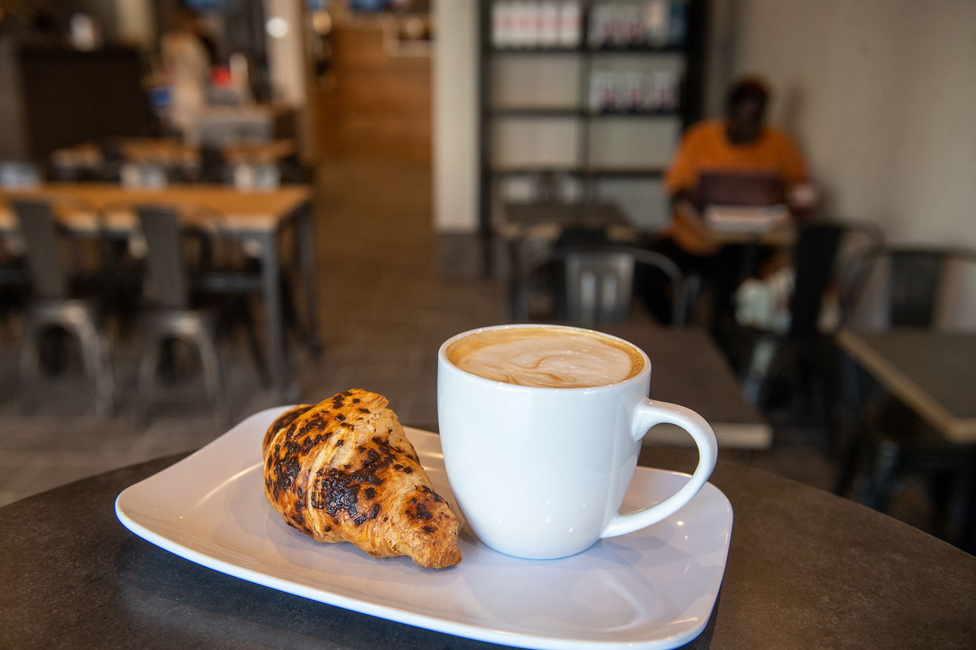 Photo: A chocolate croissant and latte with a leaf design in its foam are shown plated on a wooden coffee table. In the blurred background, people can be seen seated and working in the coffee shop.