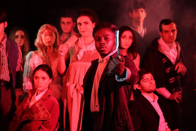 Photo of Michael Bryant Kessie (CFA’25), center, a young Black man who plays the lead role in BU On Broadways’ Sweeney Todd during a recent rehearsal. The rest of the cast stands behind him, in period attire. Everyone is lit with a deep red color.