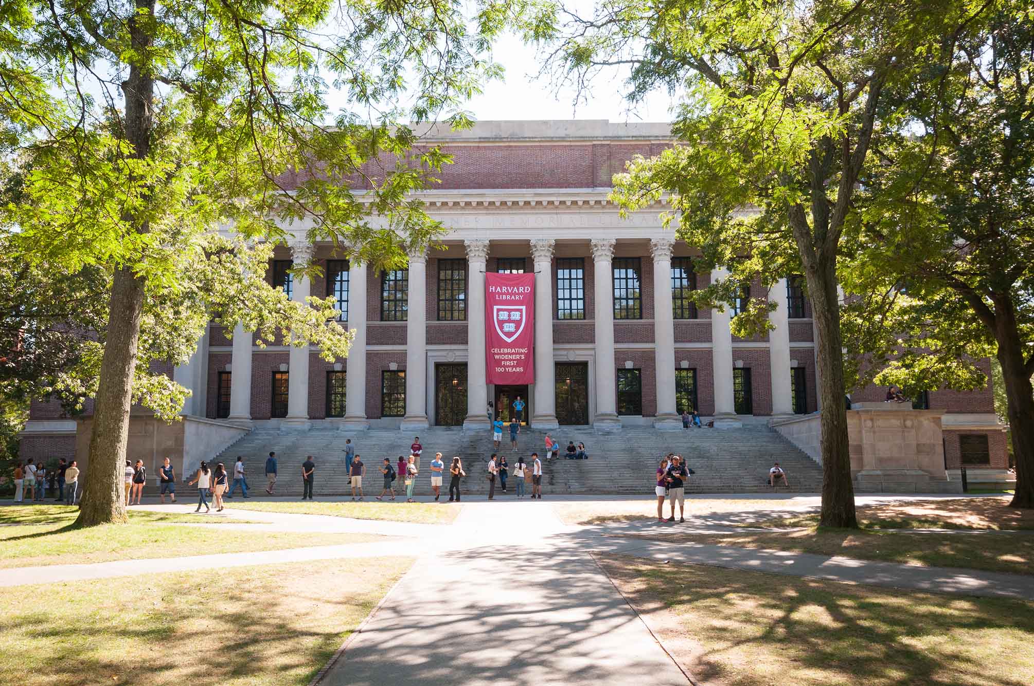 Photo: The front of a large, brick Harvard building is shown. Students walk around on campus as a large red banner with the Harvard insignia and name is displayed on the front of the building.