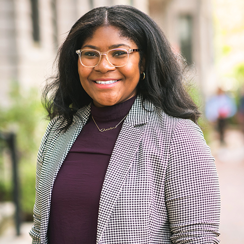 Photo of Christine Slaughter. A young Black woman wearing glasses, a burgundy sweater, and twead jacket, smiles for the camera.