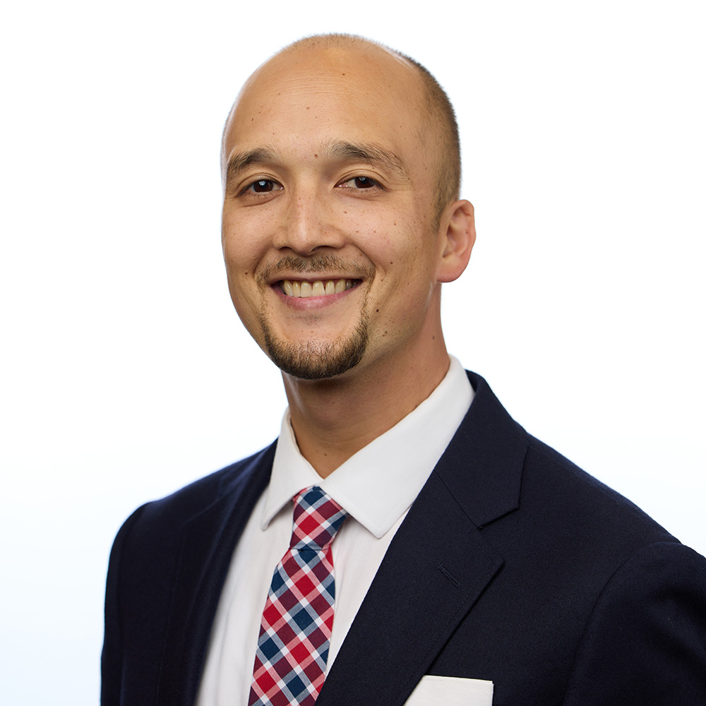 Photo: Headshot of James McCarty. A tan, bald man wearing a black suit and red and blue plaid tie smiles and poses in front of a white backdrop.