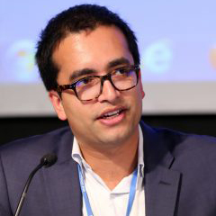 Headshot photo of Rishikesh Ram Bhandary. A Brown man wearing glasses, a white collared shirt, and navy blazer is shown as he speaks into a small microphone.