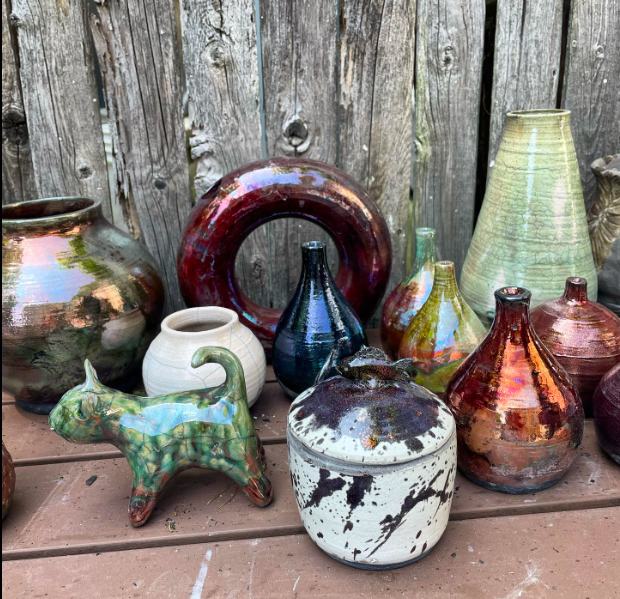 photo: Two rows of decorative, glazed pottery tumblers, vases, and figurines are displayed on a wooden table in front of a wooden wall that resembles a fence.