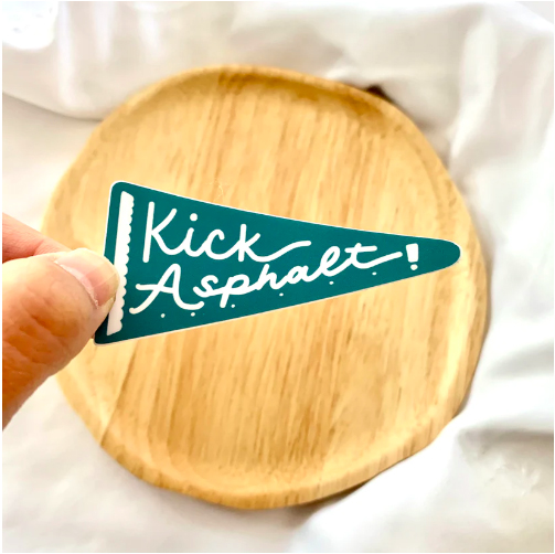 Photo: A small green flag with chalk writing that reads "Kick Asphalt!" is fancy script is shown being held by a hand. they hold the flag over a white duvet featuring a wooden plate.