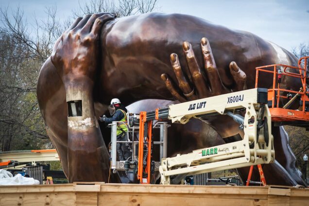 Photo: Workers install the massive bronze statue, "The Embrace" on December 6, 2022. A work works from a lowered crane on the large left hand that seems to be holding up a large bronze stone. The entire sculpture looks like two bronze hands grasping and holding up a large rock or bundle.