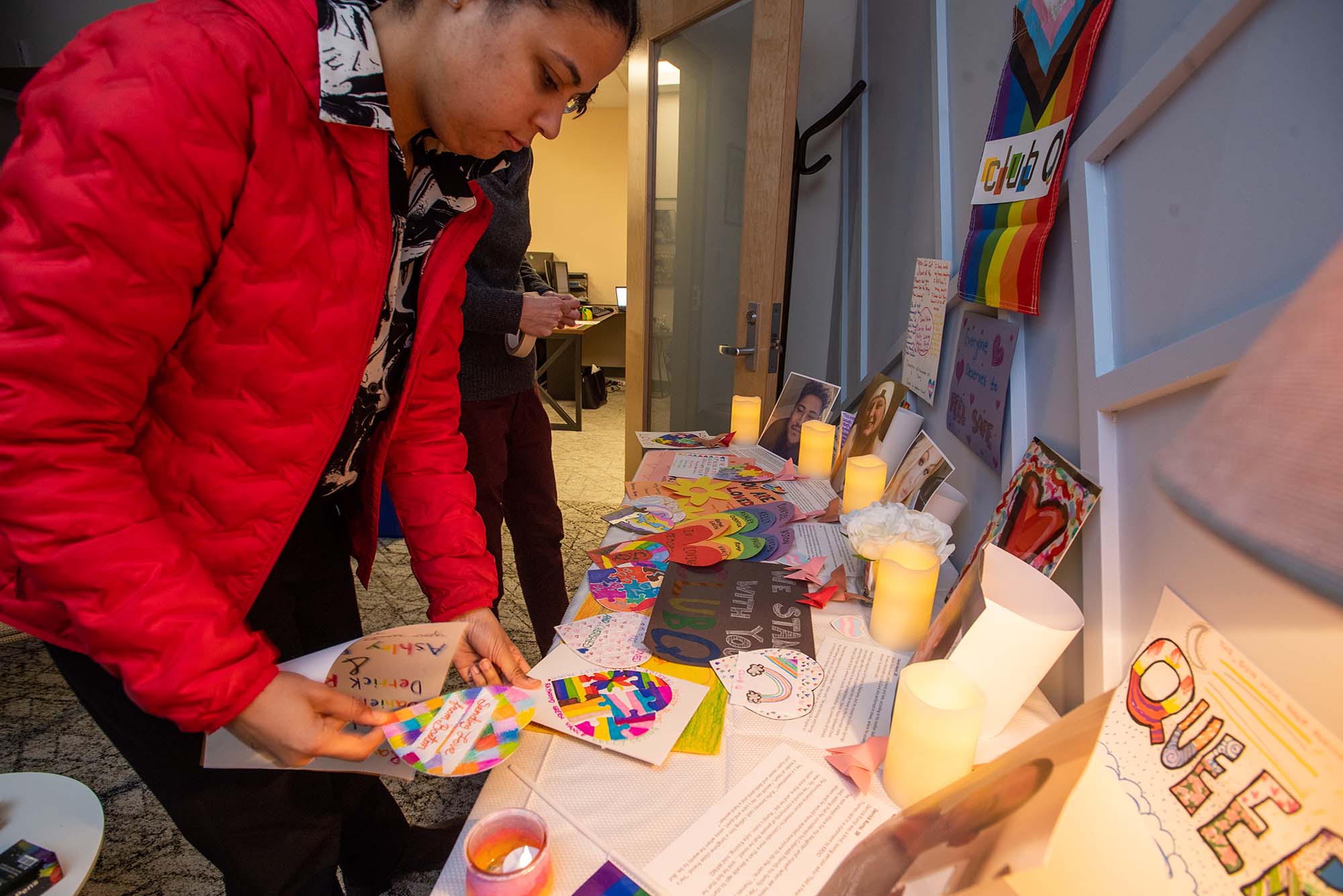 Photo: Alexis Fearing (GMS’23), programming and communications graduate assistant, foreground, and Allison Reilly, assistant director, behind Fearing, put some finishing touching on the ClubQ memorial on display in their LGBTQIA+ Center for Faculty & Staff office Dec 16. A young tan woman wearing a large red puffer coat arranges well-wishes, cards, and photos on table covered with a white table cloth.