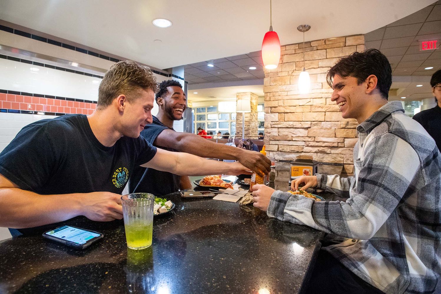Photo: (from left to right) Michael Quinn, Tim Uzoegbu, and Jordan Tab at BU West Dining Hall after a team lift. A young white man, a young Black man, and a young tan man sit at a black bar area as they joke around over dinner.