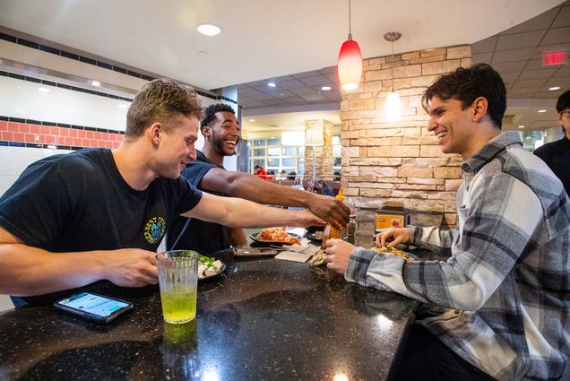 Photo: (from left to right) Michael Quinn, Tim Uzoegbu, and Jordan Tab at BU West Dining Hall after a team lift. A young white man, a young Black man, and a young tan man sit at a black bar area as they joke around over dinner.