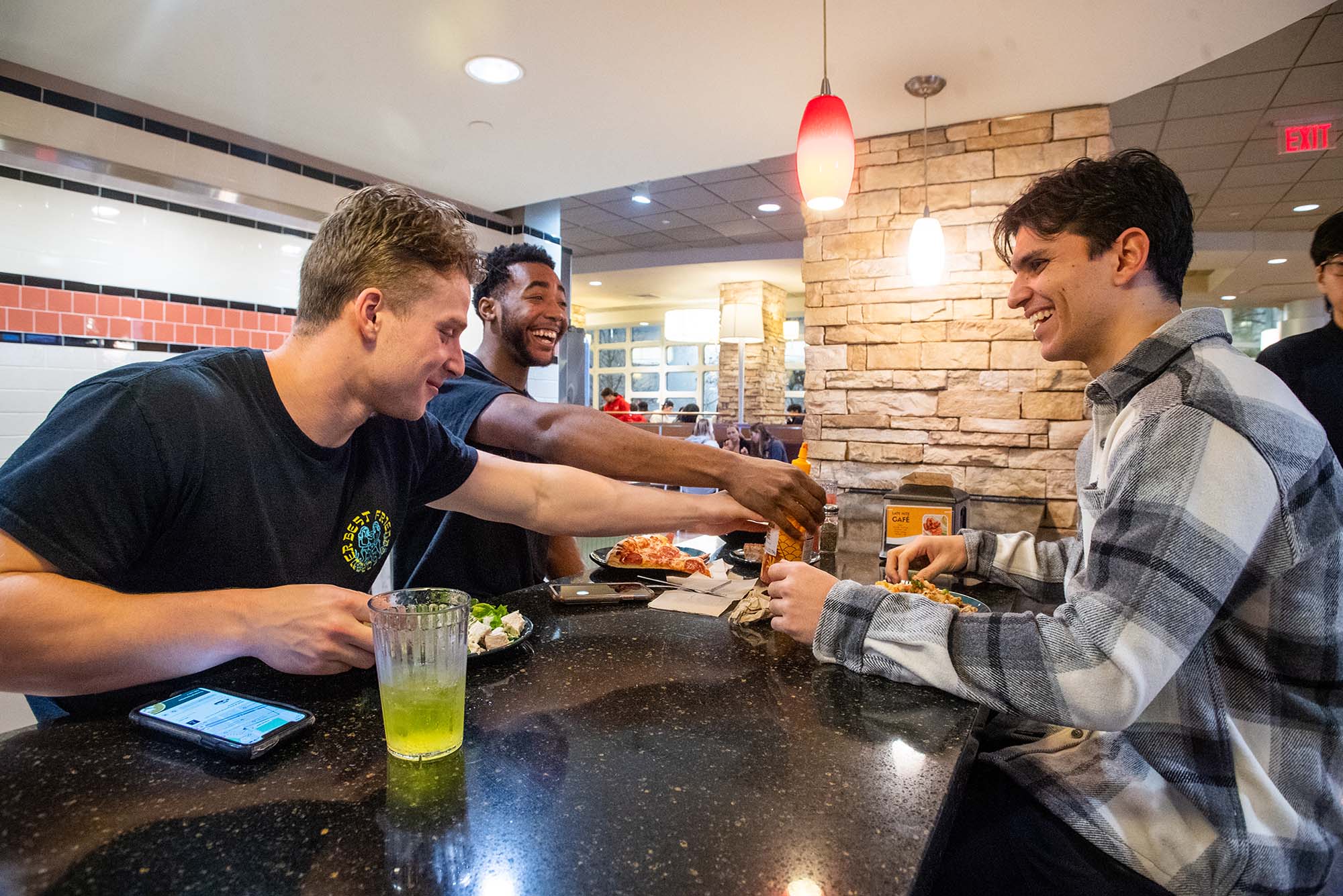 Photo: (from left to right) Michael Quinn, Tim Uzoegbu, and Jordan Tab at BU West Dining Hall after a team lift. A young white man, a young Black man, and a young tan man sit at a black bar area as they joke around over dinner.