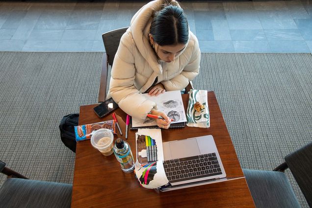 Photo: Overhead shot of Fiorella Polit (CAS’24) studying for her neuropsychology exam on the fifth floor of LAW Dec 5. A young woman in a white puffy jacket writes in a notebook in front of an open Apple laptop. Various snacks and an open colored pencil case sit around her on the table she writes on.